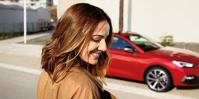 mujer sonriendo cerca de un SEAT León rojo, se destacan la flexibilidad y comodidad de los vehículos híbridos en la vida cotidiana