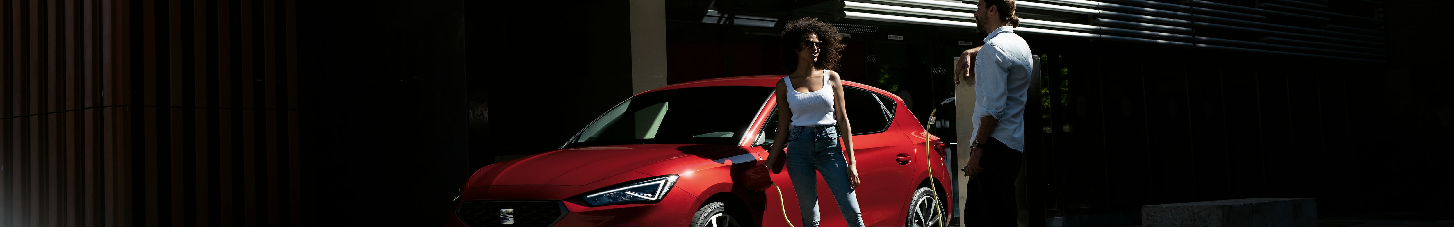 seat leon híbrido enchufable rojo y pareja debatiendo si merece la pena comprar un coche híbrido, vista de 3/4 contrapicado frente a un árbol