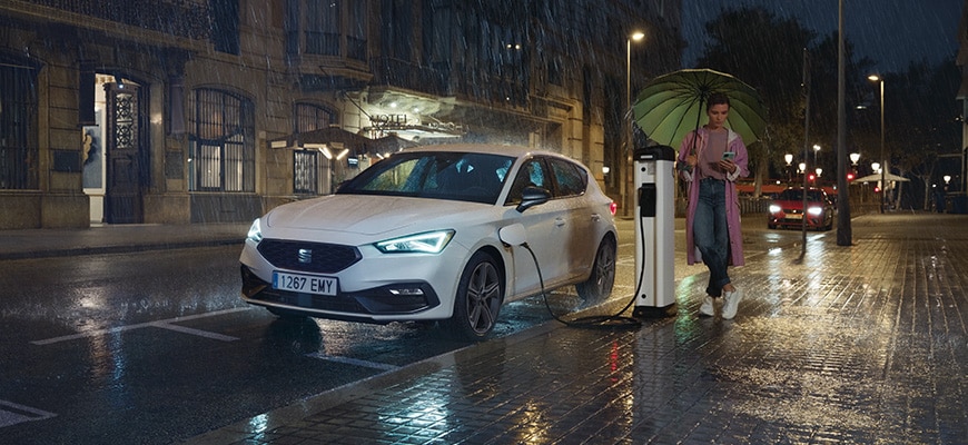 Vehículo híbrido SEAT León cargando en una estación de carga pública en una calle lluviosa por la noche, se destacan las ventajas de los vehículos híbridos